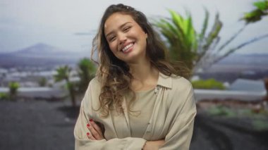 Arka planda yemyeşil ağaçları olan, kollarını kavuşturup mutluluk ve neşe saçan, açık bej bir gömlek giyen bir kadın.