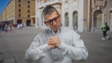 Sokakta elleri sımsıkı bağlı bir adam şehir ortamında saldırganlık gösteriyor. Gözlük ve çizgili gömlek giyiyor. Arka planda beyaz bir erkekle yüzleşiyor..