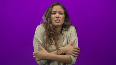 Canlı mor bir arka planda titreyen kadın, kollarını sıkıca kavuşturarak soğuk bir rahatsızlık ifade ediyor, izolasyona karşı soğuk bir güven duygusu taşıyor..