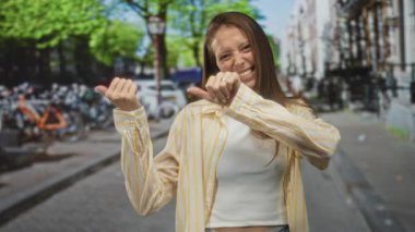 Geniş bir şekilde gülümseyen, iki baş parmağını kaldıran, sarı çizgili bir gömlek ve beyaz bir tank giyerken kalabalık bir caddede park halindeki bisikletleri ile neşeli bir güven.