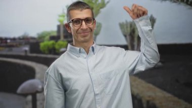 Açık havada gülümseyen adam, çevresini çevreleyen ağaçlarla dolu neşeli bir sokak ortamında olumlu ve başarıyı işaret eden başparmak kaldırıyor..