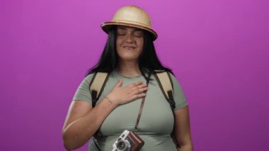 Sırt ve kaskı takmış gülümseyen bir kadın pembe bir duvarın arkasında duruyor. Kamera asılı, seyahati somutlaştırıyor ve neşe ve merakla macera yaratıyor..