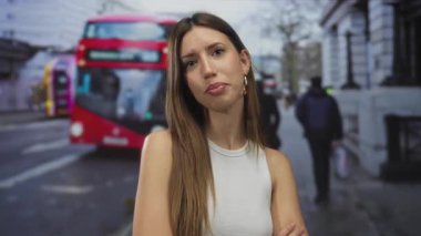 Şehir caddesinde kırmızı çift katlı otobüsün yanında kolları çapraz duran beyaz kollu ve halka küpeli bir kadın..