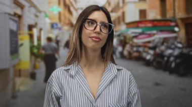 Gözlüklü, çizgili tişörtlü bir kadın mobiletler ve kafelerle dolu işlek bir sokağa bakıyor. Huzur..