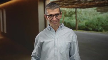 Kapalı bir odada gözlüklü, gülümseyen beyaz bir adam arka planda yeşillikle rahat bir ortamda sıradan bir görünüm sergiliyor..