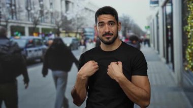 Siyah gömlekli genç adam şehir caddesinde duruyor. İnsanlar arka planda binalarla dolu kalabalık bir kaldırımda yürürken şaşkın şaşkın bakıyor..