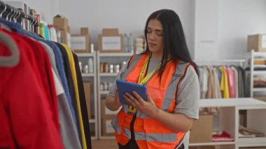 Genç bir kadın gönüllü olarak turuncu yelek giyiyor. Bağış merkezindeki kıyafetleri düzenliyor. Rafları ve rafları var..