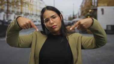 Latin kökenli genç bir kadın şehir mimarisini işaret ederken kadınlar sokakta baş parmağını gösteriyorlar..