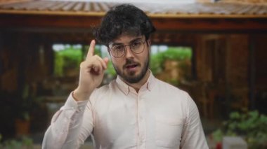 Güneşli bir günde, yeşil gözlüklü ve beyaz tişörtlü, sakallı genç bir adam, çevresi yeşilliklerle çevrili ve okuldan atılma jestini gösteriyor..