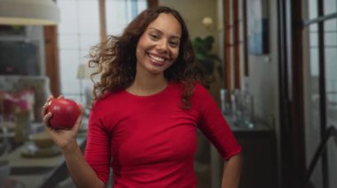 Kırmızı bluz ve altın kolye takan bir kadın, sol elinde kırmızı bir elma tutuyor, gülümsüyor ve bir binada göz kırpıyor..