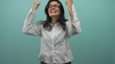 Gözlüklü ve beyaz çizgili tişörtlü bir kadın, kutlama anı için ayarlanmış nane renkli bir stüdyoda yumruklarını sıkar..