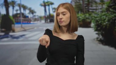 Kısa saçlı bir kadın, güneşli şehir caddesinde, bulanık palmiye ağaçlarına karşı düşünceli bir şekilde poz veriyor.