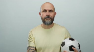 Kel sakallı adam yoğun bir ifadeyle topu beyaz arka planda tutuyor, kolunda dövmeler var..