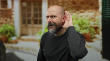Sakallı bir adam, şehir caddesinde bitki ve taş duvarla dikkatle dinliyor, günlük giysiler giyiyor ve merak gösteriyor, şehir ortamında bir açık hava sahnesini canlandırıyor..