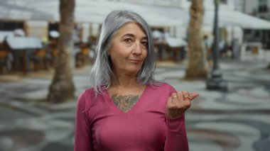 Gri saçlı ve pembe kazaklı bir kadın arka planda bulanık bir şekilde oturuyor ve ağaçlar baş parmağıyla yan tarafları işaret ediyor..