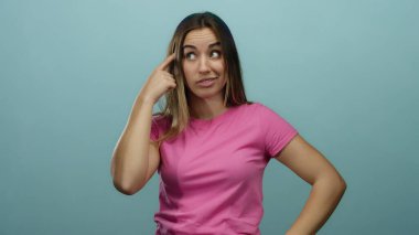Pembe tişörtlü genç kadın mavi duvara karşı şaşkın bir ifadeyle düşünüyor, yansıma ve merak uyandırıyor..