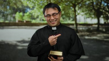 Elinde İncil 'le parkta gülümseyen Asyalı rahip, ağaçların gölgesinde siyah rahip kıyafeti giyiyor, neşe, maneviyat ve açık havada inanç taşıyor..