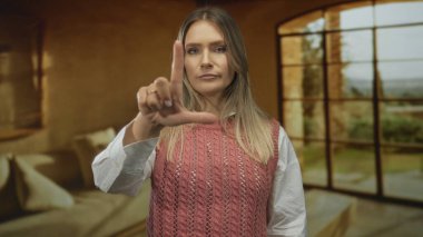 Kapalı bir otel odasında zavallı bir jest yapan bir kadın, pembe kazak giyen, sarı saçlı, geniş pencerenin yanında duran, ciddi ifade, bulanık arka plan, doğal ışık, günlük kıyafet.