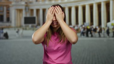 Pembe tişörtlü kadın, Vatikan şehir meydanında elleri başının üstünde, etrafı San Pedro dışından insanlarla çevrili, stres ve turizm sorunlarını yansıtıyor..