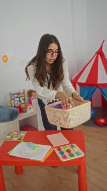 Renkli bir anaokulu sınıfında oyuncaklar düzenleyen ve etrafında eğitim malzemeleri olan bir kadın, küçük çocuklar için ilgi çekici bir öğrenme ortamı yaratıyor..