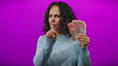 Orta yaşlı bir kadın Güney Afrika Rand banknotlarını tutuyor. Onaylamayan bir ifadeyle ve mor arka planda bir parmak hareketiyle..