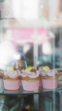 Cam bir kutuda sergilenen renkli tatlılar arasında kalabalık bir kafeterya veya fırın ortamında pembe şekerli kapkekler yer alıyor ve müşteriler için canlı ve cazip bir atmosfer sunuyor.