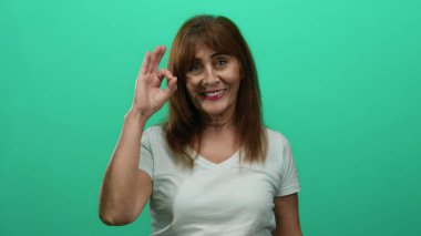 Kıdemli İspanyol kadın yeşil arka planda hoş bir jestle gülümsüyor izole bir ortamda günlük beyaz gömlek giyerek neşe ve pozitiflik sergiliyor.