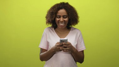 Akıllı telefon tutan ve elleri stüdyoda görülebilen limon yeşili bir fon ile gülümseyen, açık pembe tişört ve yüzük takan bir kadın..