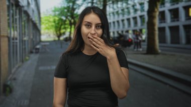 Latin kökenli genç bir kadın, şehir binaları ve ağaçların oluşturduğu kaldırım kenarlarında siyah tişört giyip, elini sandığa koydu..