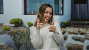 Açık bir günde, canlı bir sokak ortamını yansıtan bitkilerle dolu modern beyaz bir binanın önünde gülümseyen ve işaret eden bir kadın..