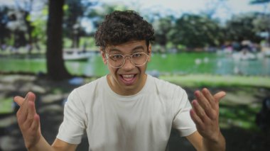 Beyaz tişört giyen ve gözlüklü genç İspanyol adam ağaçlarla dolu bir parkta yeşil bir göletin yanında konuşurken el kol hareketi yapıyor..
