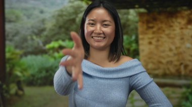 Parkta gülümseyen bir kadın, açık havada mavi bir süveter giymiş, çevresi yeşilliklerle çevrili, doğal ortamda dostça bir etkileşim ve pozitiflik sunuyor..