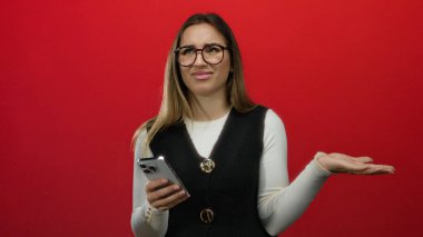Kırmızı arka planda kafası karışık bir ifadeyle akıllı telefon tutan sarışın kadın, gözlük takıyor ve günlük kıyafetler giyiyor..