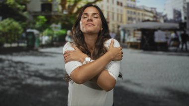 Şehir caddesinde kollarını kavuşturan bir kadın gözleri kapalı ve yumuşak bir gülümseme ile kendini kucaklıyor..