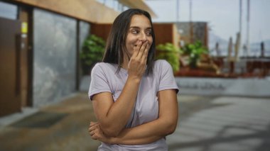 Eli ağzı kapalı bir kadın ve kollarını çapraz bina girişinde günlük tişört ve rahat duruş; çekingenlik konfor.