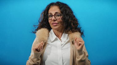 Kıvırcık saçlı, gözlüklü ve bej ceketli bir kadın, parlak mavi bir arka plana karşı poz veriyor, izole edilmiş bir stüdyo ortamında zarafet ve güven yayıyor..