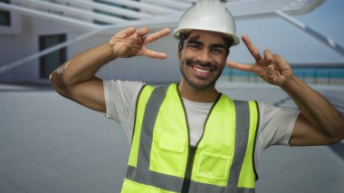 Kasketli ve yansıtıcı yelekli bir adam modern bina girişinde iki eliyle barış işareti yapıyor..