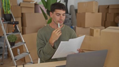 Yeşil kazaklı adam yeni ev binasında kutularla çevrili kalemle dudaklarını tutuyor, odaklanmış ifade; yeniden yerleştirme umudu planlıyor.