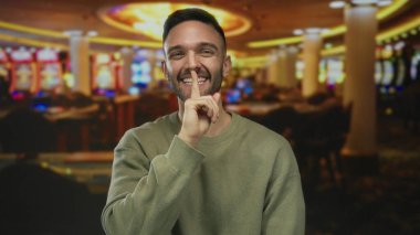 Genç adam kumarhane ortamında canlı oyunlar ve renkli ışıklarla çevrili, gülüyor ve yüzünde neşeli bir ifadeyle şşşş hareketi yapıyor..