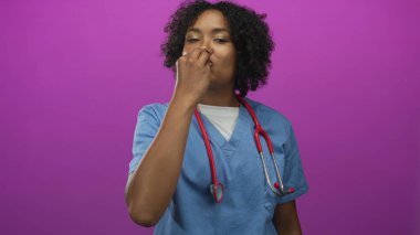 Mavi önlüklü genç Afrikalı Amerikalı kadın doktor bir eliyle parlak mor stüdyo ortamında burnunu çimdikliyor..
