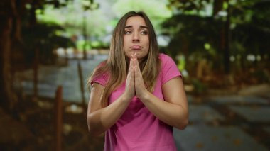 Genç bir kadın, umut dolu bir şekilde dua ediyor. Pembe gömlek giyiyor, duygularını ifade ediyor. Doğada duruyor, sakin ve düşünceli bir atmosferde..