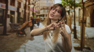 Kırmızı gözlüklü kadın, kaldırım taşı bir caddede parmakla etiket işareti yapıyor ve gülümsüyor. Bebek arabası ve banklarla görülebilir..