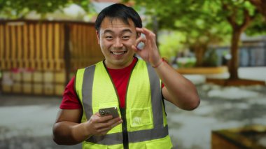 Parkta gülümseyen bir adam açık havada sarı yelek giyip akıllı telefondan mesajlaşırken iletişim ve aktiviteyi temsil ediyor..