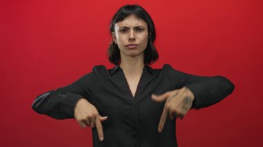 Genç İspanyol kadın parmağını sert bir ifadeyle işaret ediyor ve kırmızı stüdyoda odaklanmış bakışlarla eli dövmeli; dikkat çekme vurgusu.