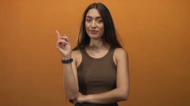 Latin kökenli genç bir kadın, turuncu stüdyo setinde kahverengi kolsuz tişört giyip yanak yanağa parmakla işaret ediyor..
