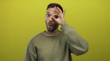 Genç İspanyol asıllı bir adam, parlak sarı bir arkaplan üzerinde el işareti yapıyor, pozitiflik ve kendine güveni gündelik kıyafetlerle ifade ediyor..