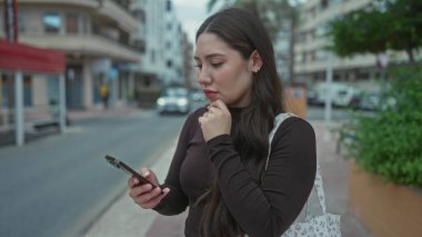 Genç İspanyol kadın akıllı telefonu tutuyor ve bir binanın yakınındaki caddede çeneye dokunuyor; yansıma iç gözlem merakı.
