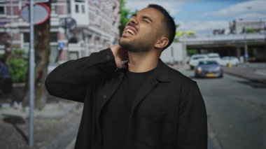 Siyah ceketli adam bulutlu gökyüzü altında şehir binalarının ortasında park halindeki arabaların yanında boynunu tutuyor;.