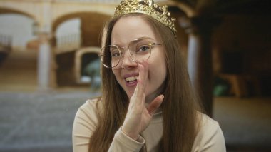 Taçlı ve gözlüklü genç beyaz kadın, antik bir binada ağzını usulca kapatıyor..