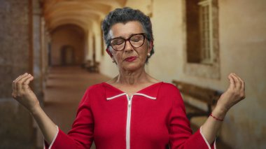 Kırmızı elbiseli yaşlı bir kadın eski bir kasaba caddesinde huzur içinde meditasyon yapıyor, sakin bir şehir ortamında sükuneti somutlaştırıyor..